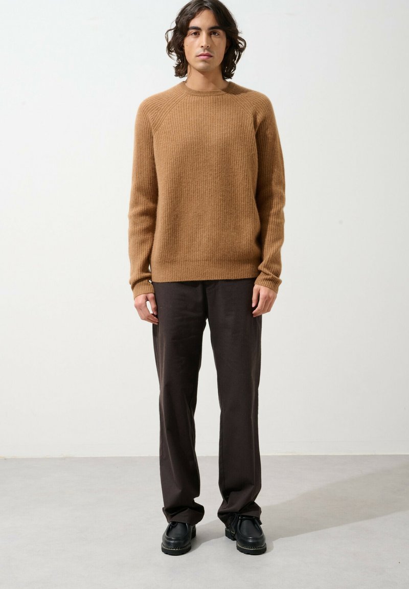 Brauner gerippter Strickpullover mit langen Ärmeln, kombiniert mit dunklen Hosen und schwarzen Schuhen, zeigt einen lässigen Schnitt und eine strukturierte Textur.