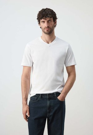 Mann mit lockigem dunklem Haar, der ein schlichtes weißes T-Shirt mit kurzem V-Ausschnitt und dunkelblaue Jeans trägt, mit einer Hand in der Tasche.
