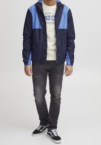 Coupe-vent bleu marine et bleu clair avec capuche, fermeture éclair, poignets élastiques, et un sweat à capuche gris foncé en dessous ; associé à un jean noir et des baskets.