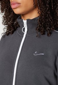 Šedá zipová mikina Nike s bílým zipem, žebrovaným límcem a diskrétním logem na hrudi. Látka vypadá hladce s čistým designem.