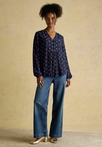 Plūstoša tumši zilā blūze ar ziedu rakstu, V veida kakla izgriezumu un garām piedurknēm, apvienota ar plašām džinsu biksēm un zelta bloku papēža apaviem.