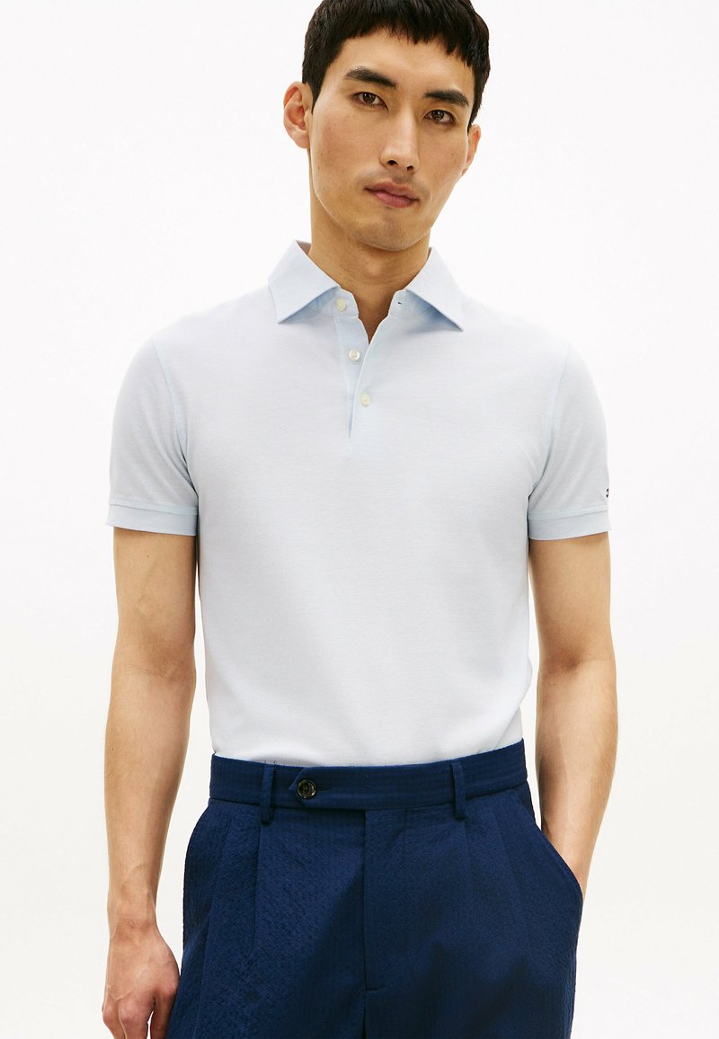 Homme portant un polo à manches courtes bleu clair, rentré dans un pantalon taille haute texturé bleu foncé avec une fermeture à bouton.