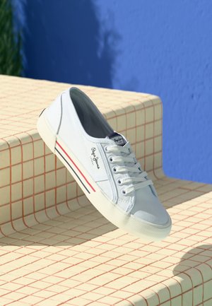 White low-top sneaker with laces and red and blue stripes on the sole, displayed on checkered beige steps against a blue wall.