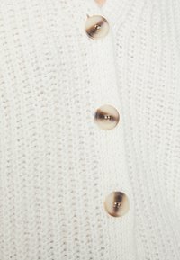 Vit stickad kofta med mönstrad design, försedda med tre runda ljusbruna knappar framtill.