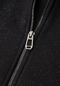 Czarne zbliżenie na materiał z zamkiem błyskawicznym o błyszczącej fakturze, z srebrnym zamkiem i napisem "STREET ONE" na suwaku.