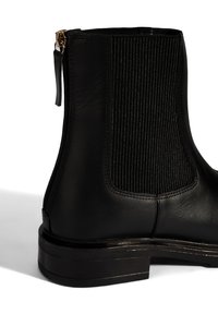 Schwarze Lederstiefelette mit gerippter elastischer Seitenpartie, niedrigem Blockabsatz und goldenem Reißverschluss-Zugband auf weißem Hintergrund.
