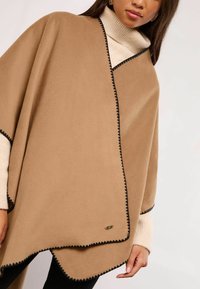 Dzeltena flīsa pončo ar melnu viļņu apmalēm un zelta detaļu. Mīksta tekstūra, atvērta dizaina, uzlikta uz krēmkrāsas džempera.