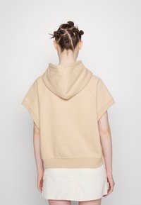 Beige zkrácená mikina s kapucí, krátkými rukávy a hladkým povrchem. Má ribbované zakončení a uvolněný střih. Zobrazená zadní strana.