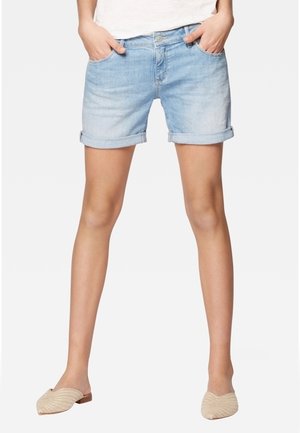 Person iført lyseblå denimshorts, hvid top og beige flettede ballerinaer står med hænderne i lommerne mod en ensfarvet baggrund.