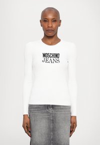 Bela rebrasta dolga majica z dolgimi rukavi, ki vsebuje črn napis "Moschino Jeans". Skupaj zivo v kombinaciji s sivo denimsko krilom za popoln izgled.