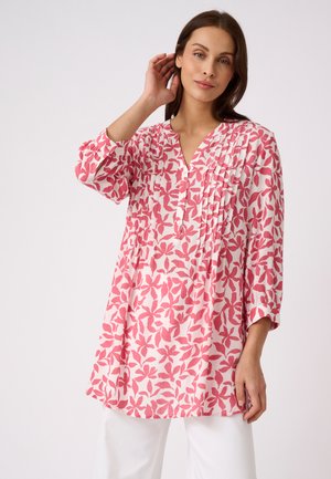 Donna che indossa una tunica a maniche lunghe con motivo floreale rosa e bianco con bottoni e pantaloni bianchi, posando con una mano vicino alla testa.