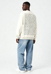 Šviesiai kreminės spalvos megztinis su juodu tekstu ant nugaros, derinamas su laisvo kirpimo mėlynais džinsais ir baltais sportbačiais, prieš neutralų foną.