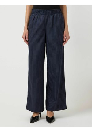 Kvinne iført marineblå vidbente bukser med striper, elastisk midje og svarte spisse hæler mot en ensfarget bakgrunn.