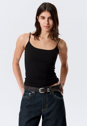 Jeune femme aux cheveux bruns mi-longs portant un haut noir à fines bretelles et un jean bleu foncé avec une ceinture noire, les mains dans les poches.