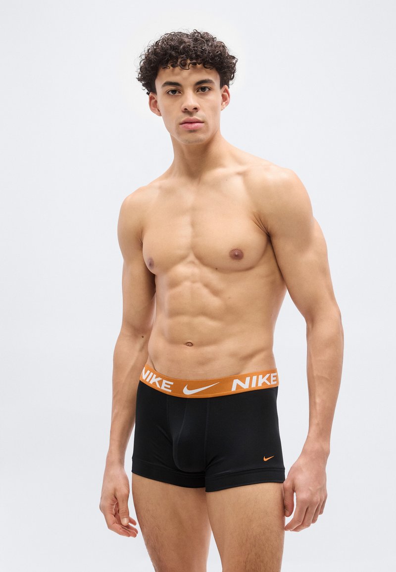 Μυώδης νεαρός με σγουρά μαλλιά που φοράει μαύρα boxer briefs της Nike με πορτοκαλί ζώνη μέσης και λογότυπο, στέκεται μπροστά σε απλό φωτεινό φόντο.