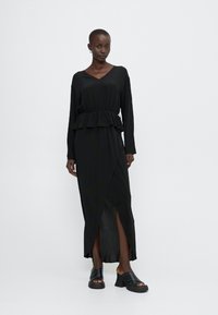 Musta pitkähiuniformupuku, jossa on peplum-vyötärö, kietaisu-design ja sivureikä. Valmistettu sileästä kankaasta, käytetty paksujen mustien kenkien kanssa.