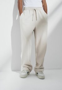 Bežīgas sporta bikses ar elastīgu jostu un auklu, ar sānu kabatām un brīvu piegriezumu, kopā ar baltām un zaļām sporta kedām.