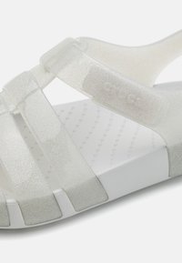 Průhledné, třpytivé sandály na pásku ve světle šedé barvě s texturovanou bílou stélkou a tvarovanou podporou klenby. Obsahuje logo na pásku.