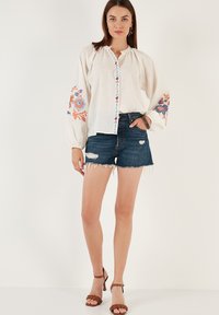 White embroidered blouse with long sleeves, featuring colorful floral patterns on cuffs, paired with distressed denim shorts and brown heeled sandals.