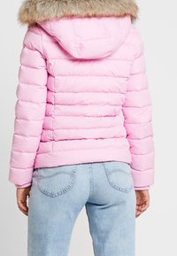 Puffermantel in Hellrosa mit horizontalem Steppmuster, ausgestattet mit einer mit Fell gefütterten Kapuze und Strickbündchen. Getragen mit hellblauen Jeans.