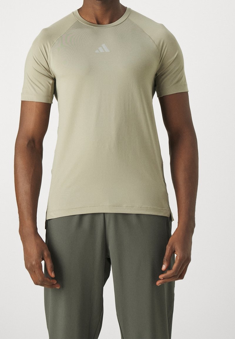 Camiseta deportiva de color verde oliva con mangas cortas, cuello redondo y un logo reflectante en el pecho. Tejido suave y aberturas laterales.