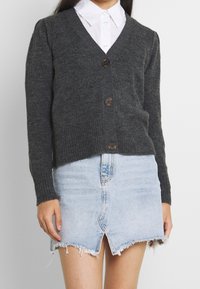 Grau gestrickte Strickjacke mit V-Ausschnitt, fünf Knöpfen und geripptem Saum, kombiniert mit einem hellblauen Jeansrock mit ausgefranstem Rand und Schlitz.