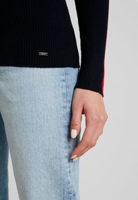 Main reposant à côté d'un jean en denim bleu clair, portant un pull côtelé noir avec une bande rouge sur la manche et une petite étiquette logo.