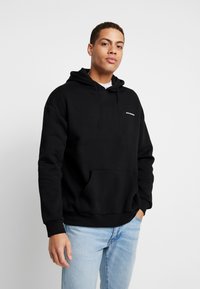 Black hooded sweatshirt made from cotton fabric, featuring a front pocket and small white text logo on the left side, with ribbed cuffs.