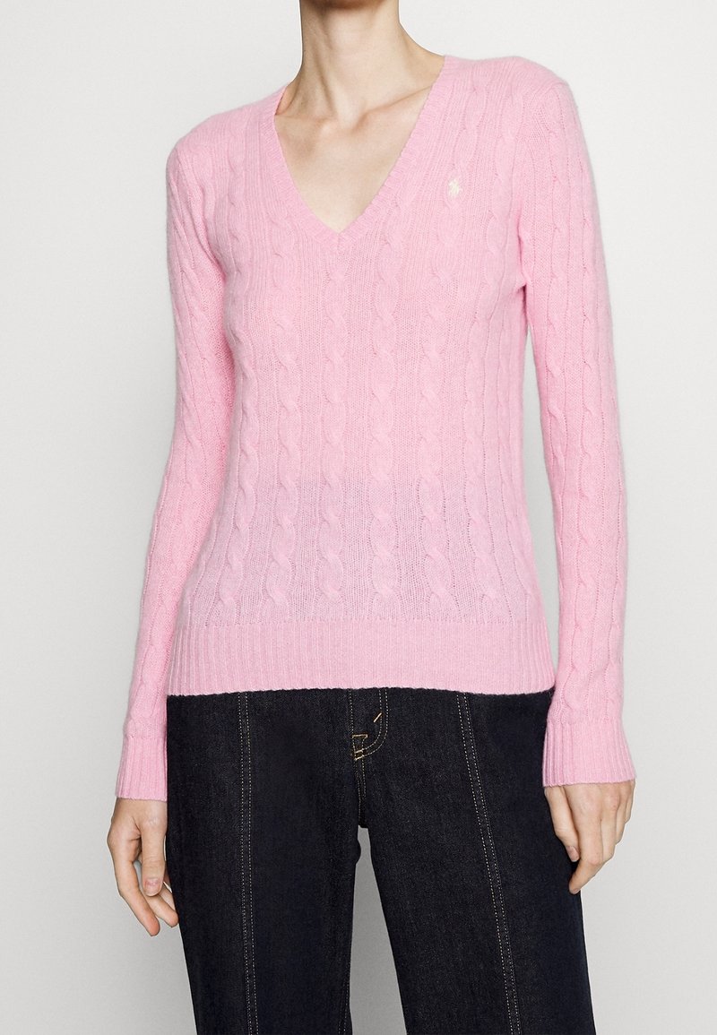 Pull rose clair à col en V en maille torsadée porté avec un jean bleu foncé, montrant le haut du torse et les bras d'une personne debout.