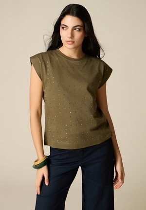 Donna con lunghi capelli scuri che indossa un top senza maniche color verde oliva con borchie, pantaloni blu navy e braccialetti spessi su uno sfondo beige.