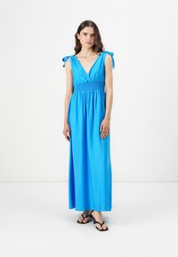 Maxi kjole i en lys blå stof med V-hals, smocket talje og skulderremme. Sammen med sorte flip-flops med glat overflade.