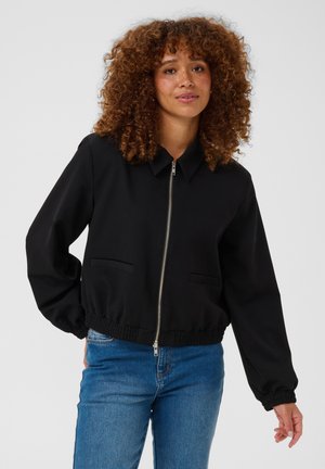 Femme aux cheveux bouclés portant une veste noire zippée et un jean bleu, debout devant un fond clair uni.