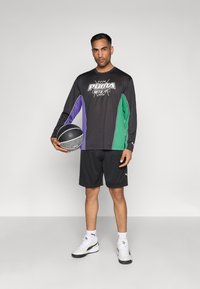 Μαύρο μακρυμάνικο πουκάμισο με μωβ και πράσινες λεπτομέρειες, με λογότυπο Puma. Συνδυάζεται με μαύρα σορτς και λευκά sneakers, κρατώντας μια μπάλα μπάσκετ.