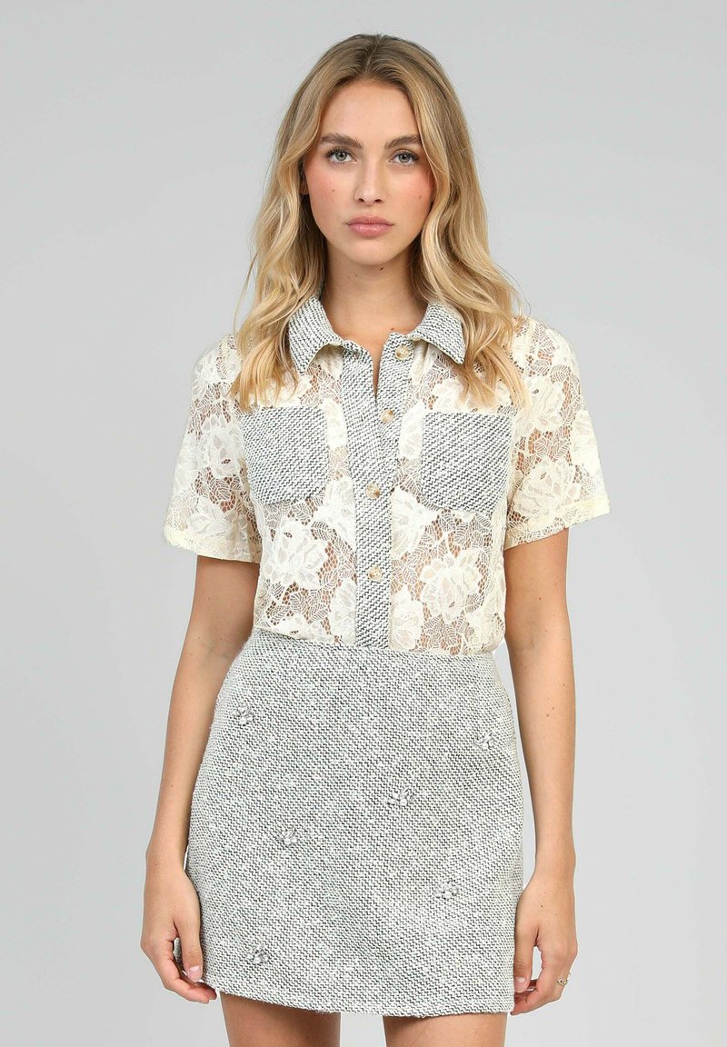 Blouse en dentelle à manches courtes et jupe grise texturée ; comporte deux poches poitrine et une fermeture à boutons ; motif en dentelle florale complexe.