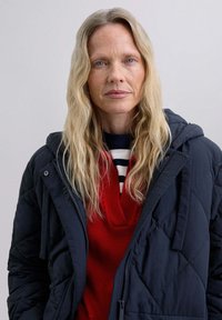 Quilted navy jacket with hood, layered over a red knit sweater featuring a V-neck and horizontal striped details. Long, wavy blonde hair.