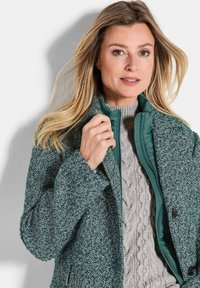 Femme aux cheveux blonds portant un manteau vert texturé sur un pull en tricot torsadé gris clair, ajustant le col du manteau.
