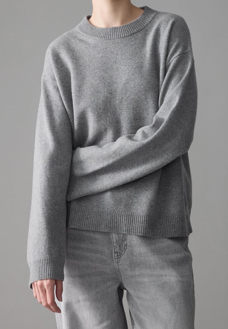 Personne portant un pull en maille gris avec poignets et ourlet côtelés, associé à un jean gris clair, debout devant un fond gris uni.