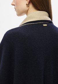 Παλτό navy από μείγμα μαλλιού με αντιθέσιμο μπεζ γιακά και διακριτική χρυσή λ�επτομέρεια λογότυπου· διαθέτει κομψή σιλουέτα και υφασμάτινη υφή.