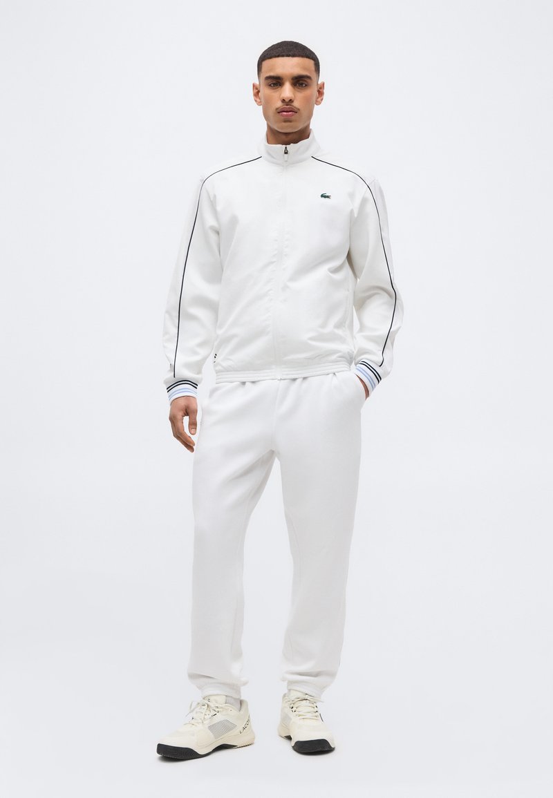 Νεαρός άνδρας που στέκεται, φορώντας λευκό αθλητικό μπουφάν με μαύρες γραμμές, λευκό παντελόνι που ταιριάζει και εκρού αθλητικά παπούτσια με μαύρες σόλες.