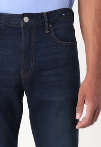 Dark blue denim jeans with visible stitching details in orange, front pockets, and a metal button closure. Smooth texture and fitted design.