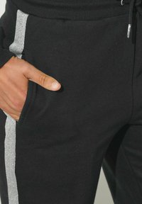 Schwarze Jogginghose mit einem silbernen Glitzerstreifen an der Seite, im bequemen Schnitt und mit einer Vordertasche. Weicher Stoff, lässiger Stil.
