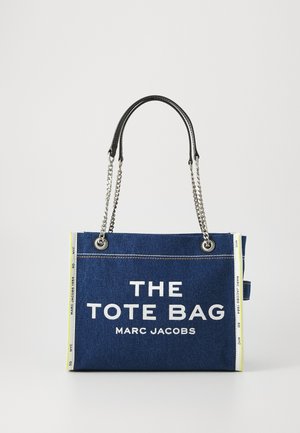 Bolso tote de denim con texto blanco "THE TOTE BAG MARC JACOBS" en el frente, asas de cuero negras, acentos de cadena plateada y un detalle amarillo en contraste.