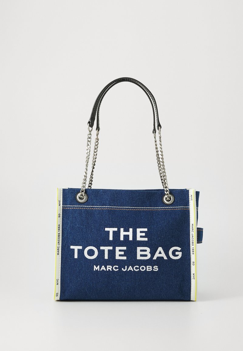 Džinsa soma ar baltu uzrakstu "THE TOTE BAG MARC JACOBS" priekšpusē, melnām ādas rokturēm, sudraba ķēdes akcentiem un kontrastējošu dzeltenu detaļu.
