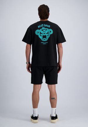 Man draagt zwarte T-shirt met turquoise abstract gezicht ontwerp en tekst, staat met rug naar camera op effen achtergrond.