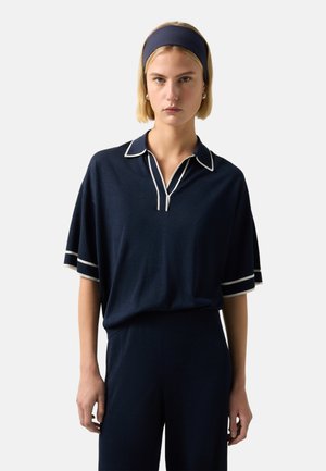 Kvinde iført en marineblå skjorte med hvide kanter og matchende bukser, accessoriseret med en bred navy hårbøjle og creol øreringe.