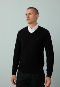 Schwarzer Strickpullover mit V-Ausschnitt, strukturiertem Muster und Rippbündchen. Enthält ein kleines Logo auf der Brust. Trägt man über einem weißen Hemd mit Kragen.