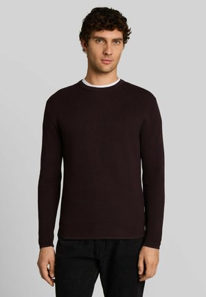 Jonge man met krullend donker haar die een donker bordeauxrode trui met lange mouwen en zwarte broek draagt, staand tegen een effen lichtgrijze achtergrond.