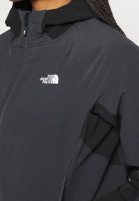 Sort hættejakke med høj krave, lavet af tekstureret stof. Har lynlås og et hvidt logo på brystet.