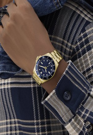 Wrist wearing a gold metal watch with a black face showing 10:09, over a blue and beige plaid shirt cuff with button detail.