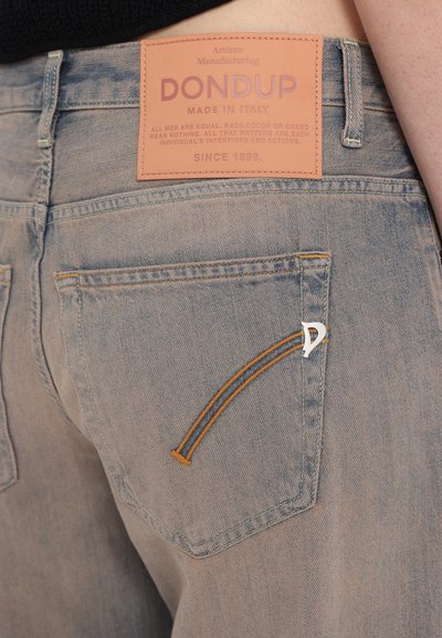 Jeans de ganga de um azul claro com um detalhe de bolso em laranja costurado e uma etiqueta em pele bege com marcação em relevo na cinta.